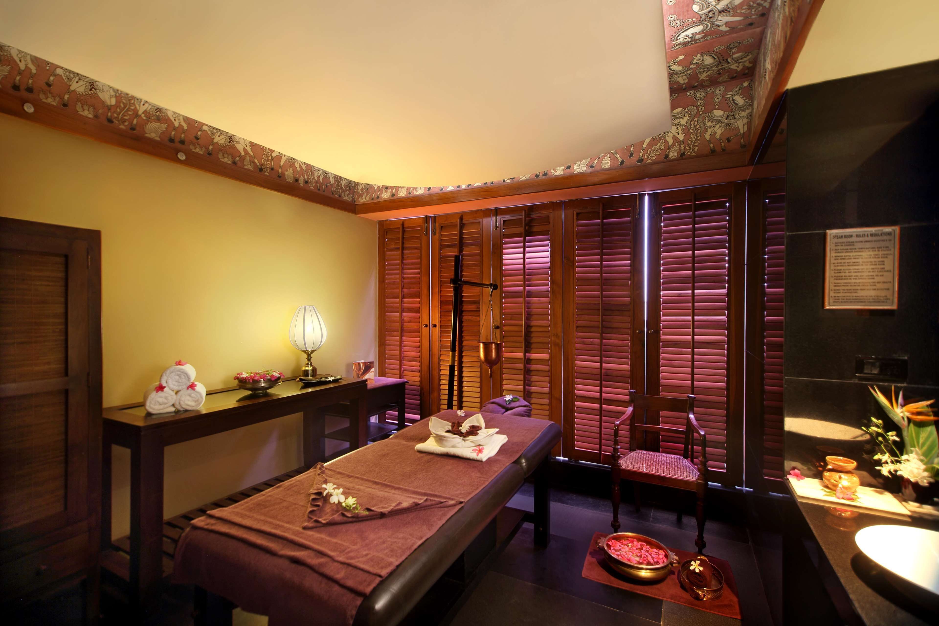 A massage room with a wooden massage table, a chair, a small table with towels and a lamp, and a sliding door with patterned curtains.