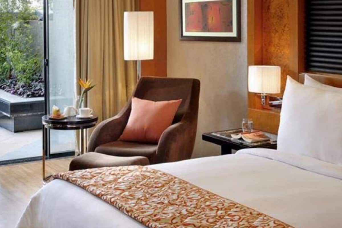 Una habitación de hotel con cama, sillón, mesa, lámpara y un cuadro en la pared.