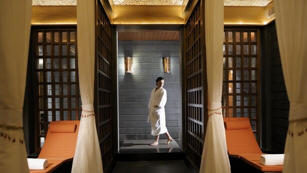 Habitación de spa con una persona en bata, dos sillones de masaje y una pared oscura con un patrón de cuadrícula de madera.