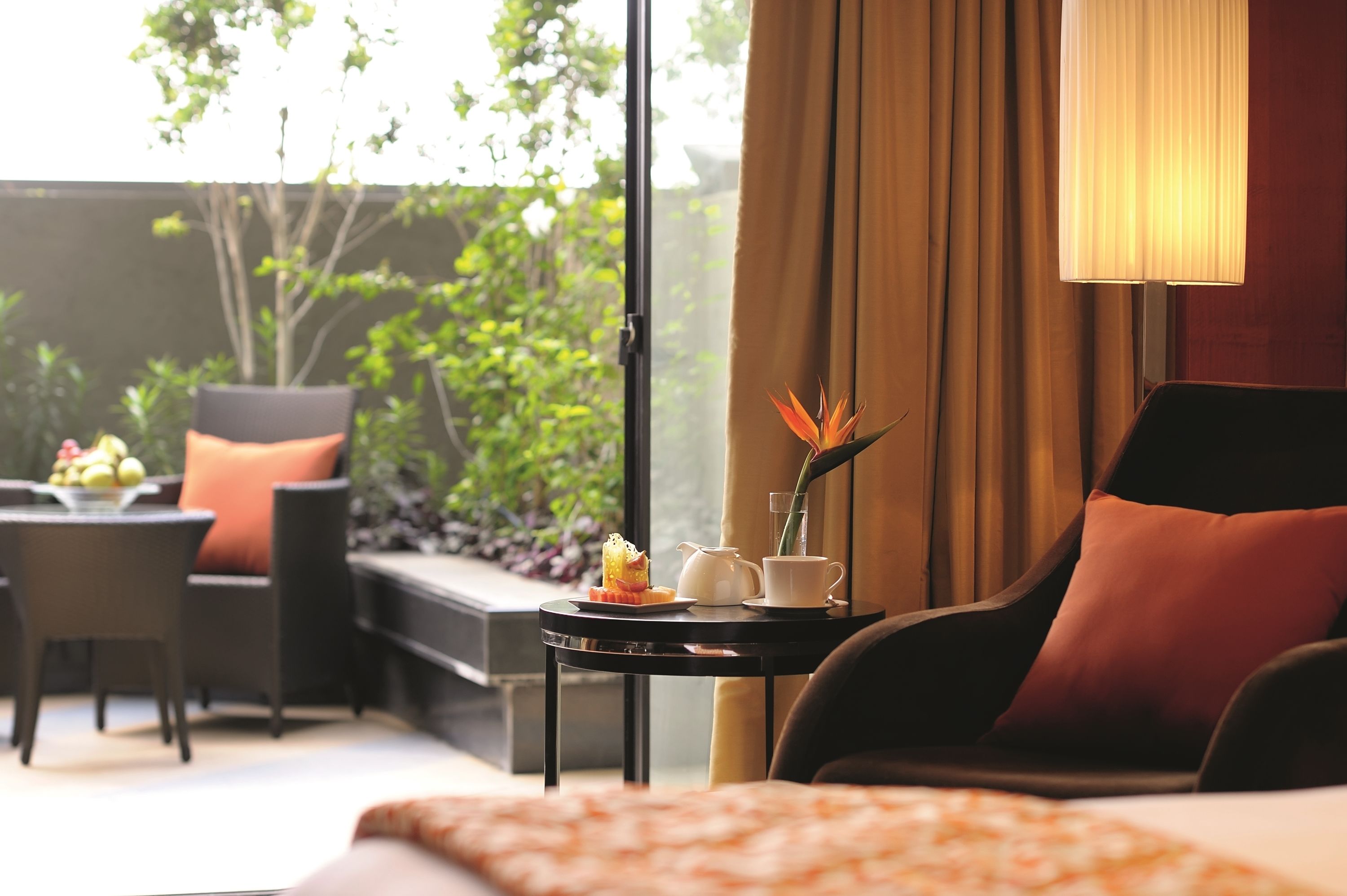 Ein Hotelzimmer mit einem Bett, einem Sessel, einem kleinen Tisch mit Lampe, einer Vase mit Blume und Blick ins Grüne.