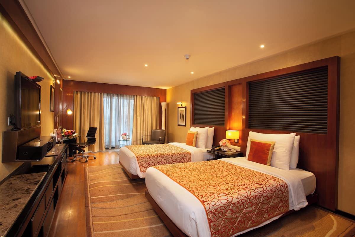 A hotel room with two beds, a desk, a chair, a TV, and a window with curtains.