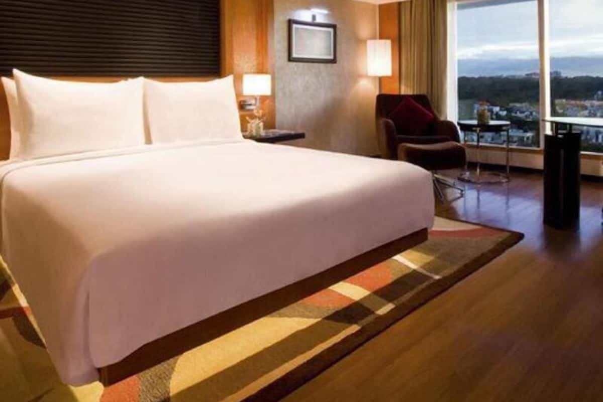 A hotel room with a large bed, a desk, a chair, and a view of the city through large windows.