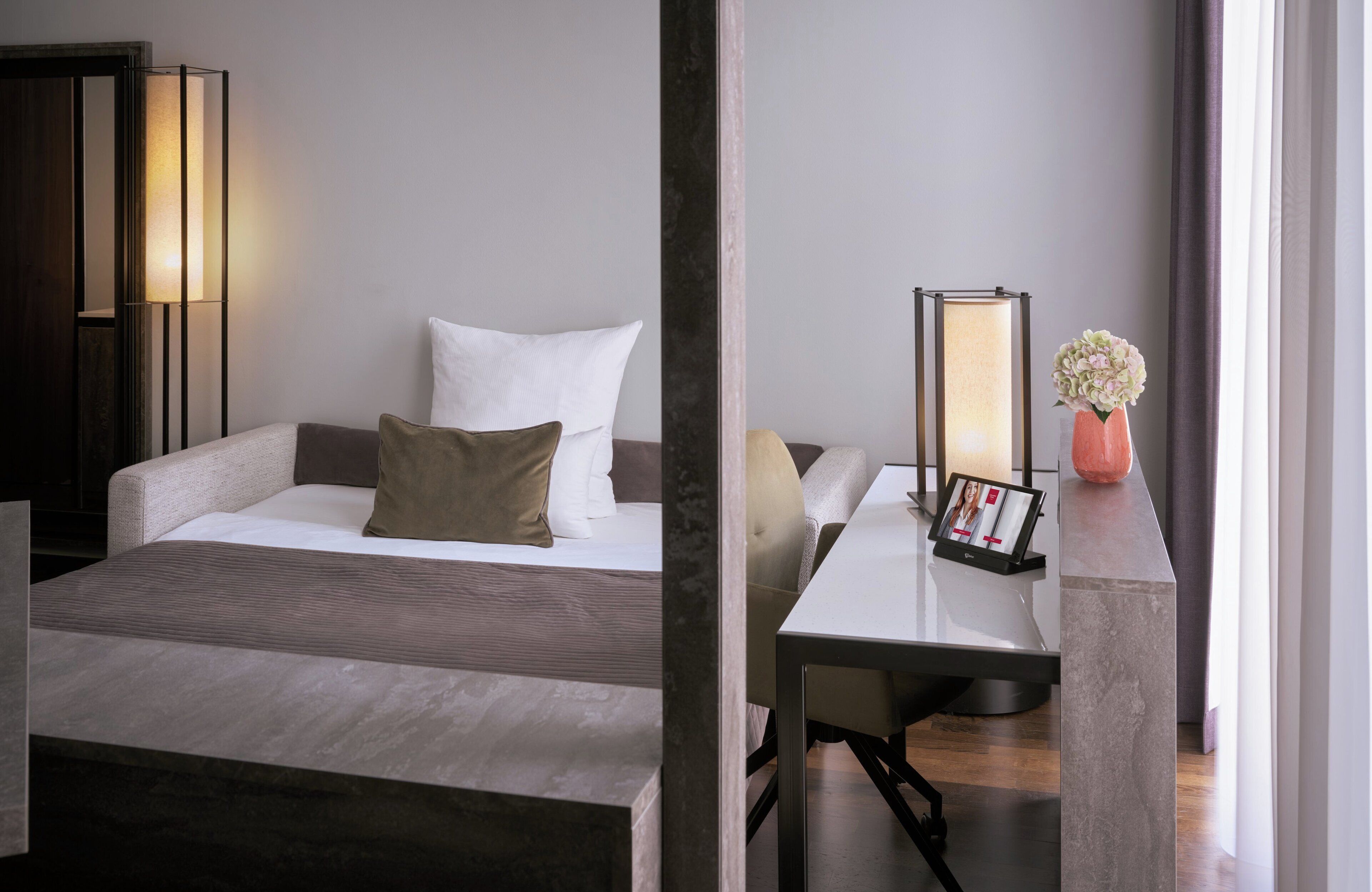 Ein modernes Hotelzimmer mit Bett, Nachttisch, Lampe und einer Vase mit Blumen.