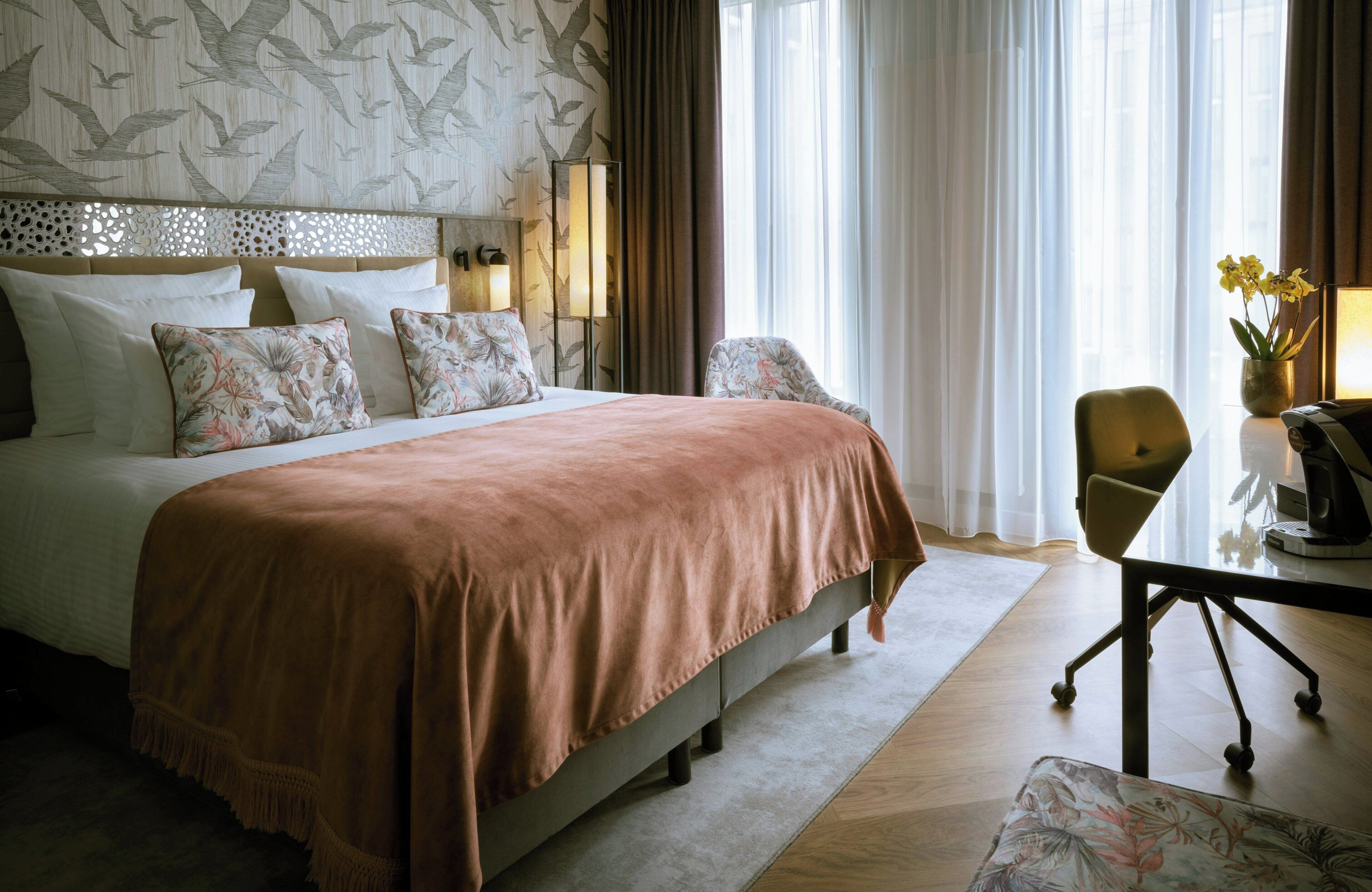 Habitación de hotel con una cama grande, un escritorio con silla, un jarrón con flores y una ventana con cortinas.