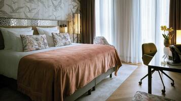Habitación de hotel con una cama grande, un escritorio con silla, un jarrón con flores y una ventana con cortinas.