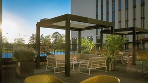 An outdoor patio area with seating arrangements, a pool, and a modern building in the background. - Leonardo Royal Hotel Munich (Munich)