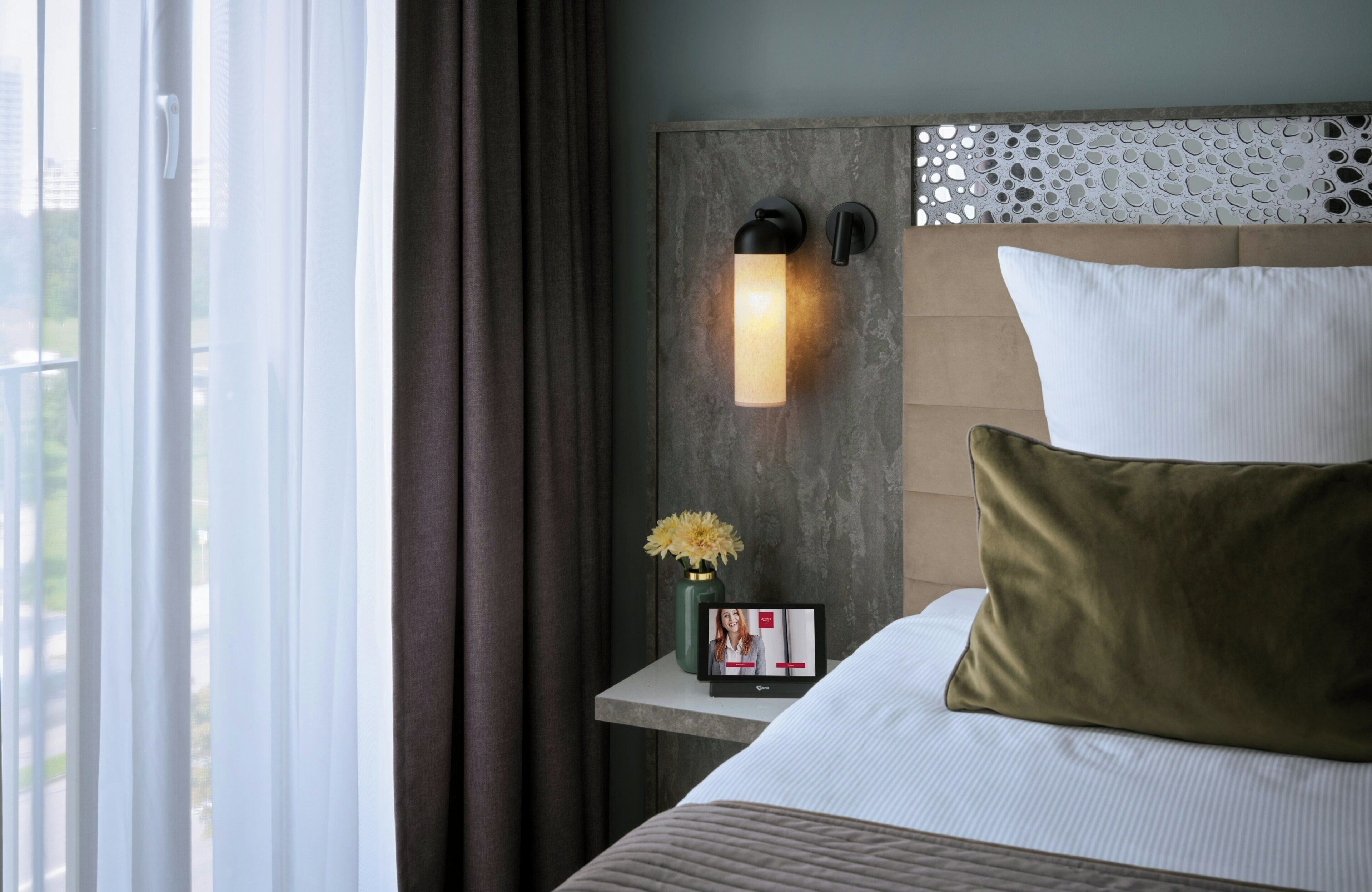A hotel room with a bed, a nightstand with a vase of flowers, and a wall-mounted light fixture.