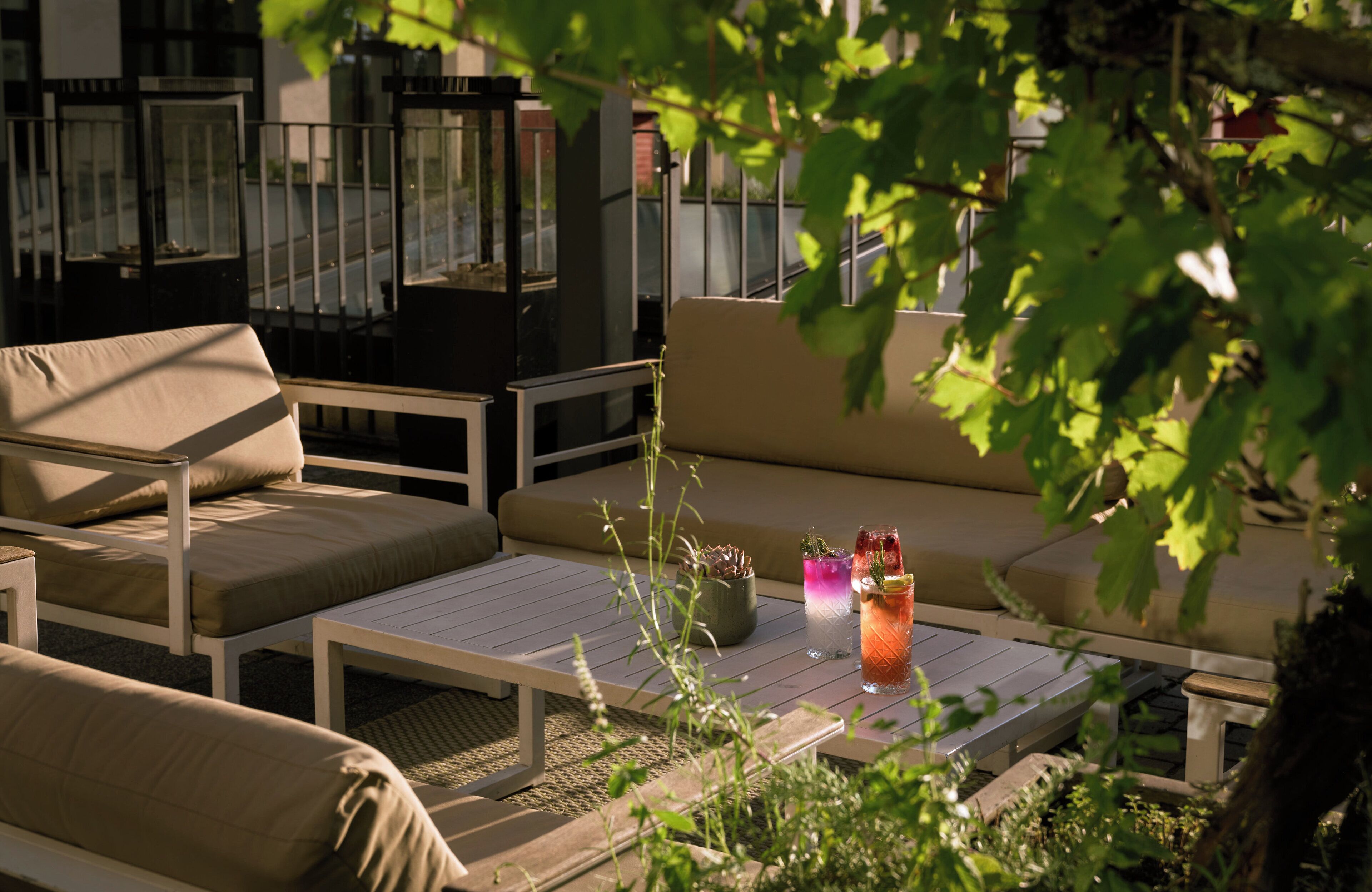 A sunlit outdoor seating area with cushioned sofas and a table, featuring a drink and a potted plant.