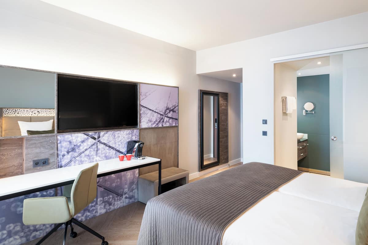 A modern hotel room with a large bed, a desk with a chair, a television, and a bathroom with a mirror and sink.