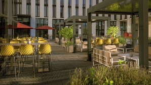 An outdoor cafe area with yellow chairs and tables, surrounded by a modern building with large windows. - Leonardo Royal Hotel Munich (Munich)