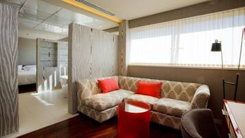 A modern hotel room with a sofa, a red ottoman, and a bed with a white headboard.