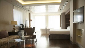 Habitación de hotel con cama, sofá, escritorio con lámpara, televisor y baño a la vista a través de una puerta abierta.