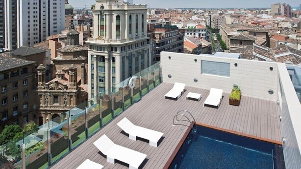 Un área de piscina en la azotea con sillones de descanso y una barandilla de cristal con vistas a un paisaje urbano.