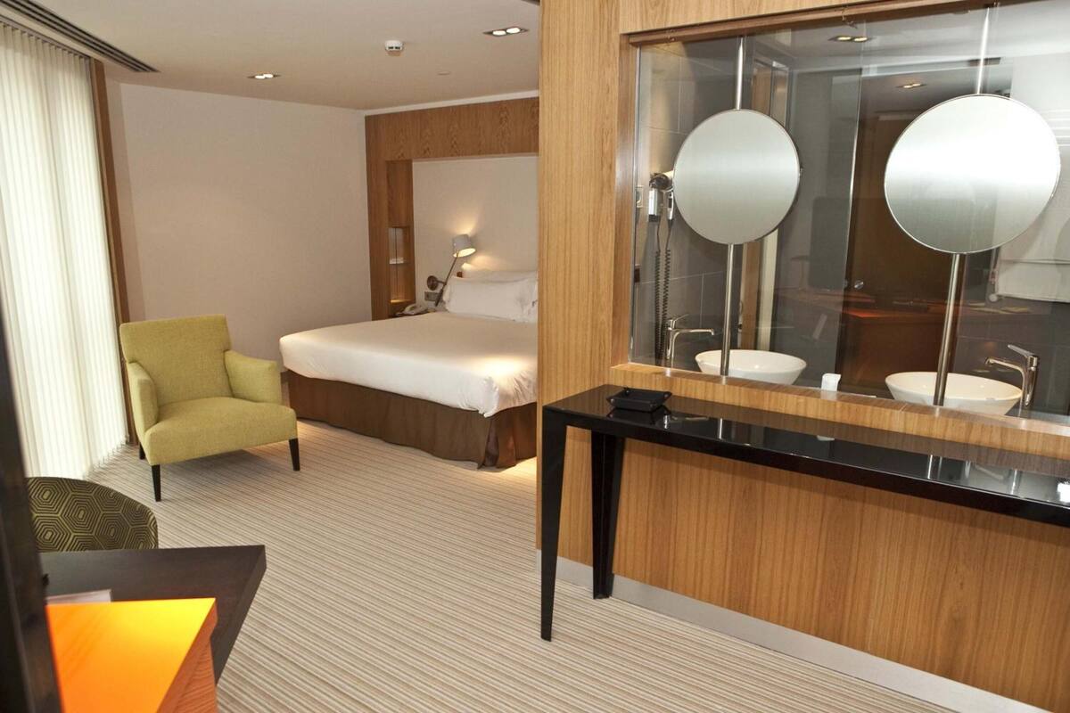 A hotel room with a bed, a sitting area with a chair, a vanity with a mirror, and a desk.