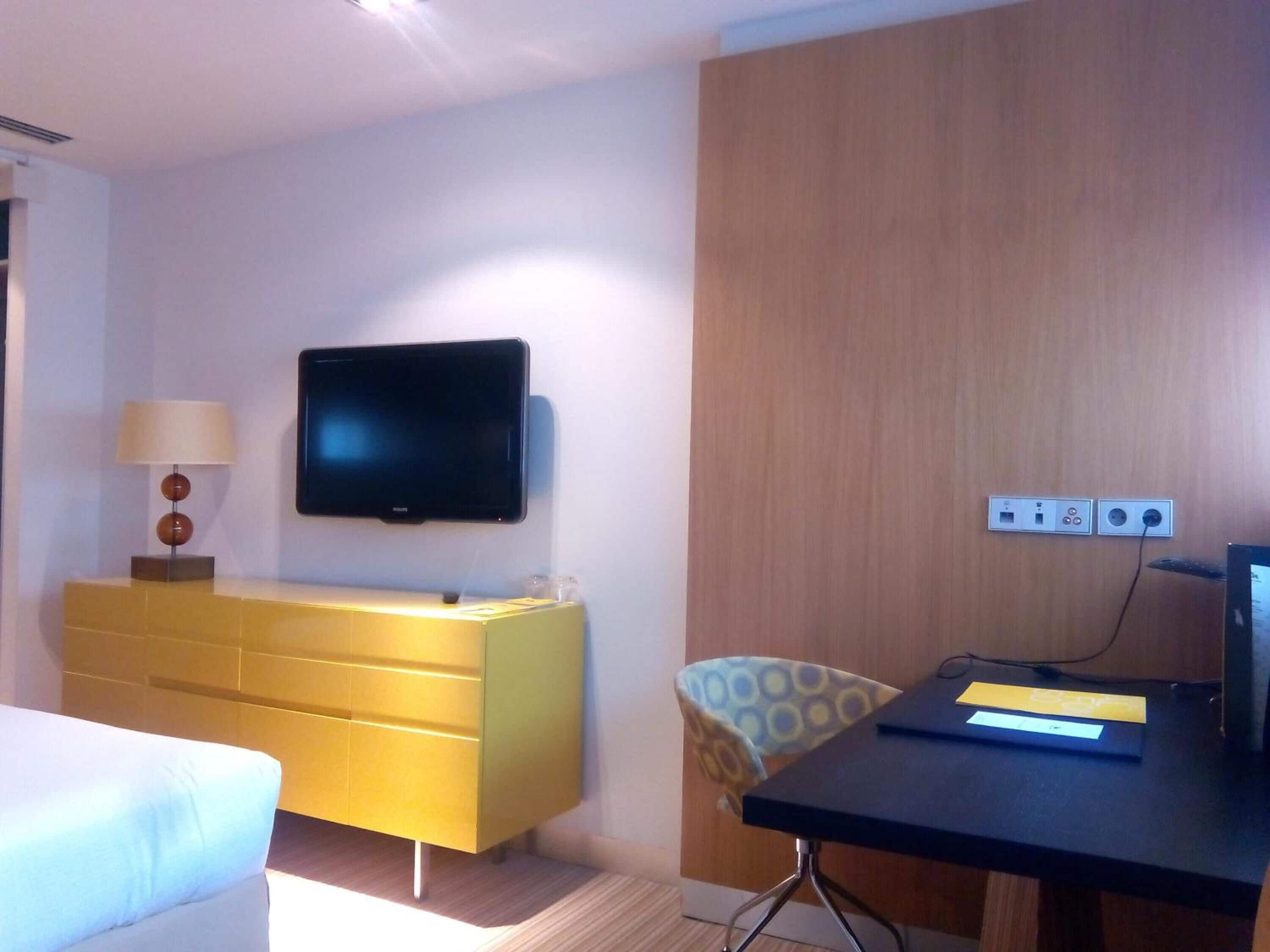 A hotel room with a flat-screen TV mounted on the wall, a yellow dresser, a desk with a computer, and a patterned chair.