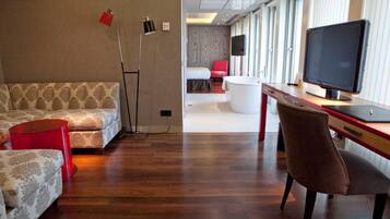 Una habitación de hotel moderna con suelo de madera, un sofá, un escritorio con ordenador y una silla roja.