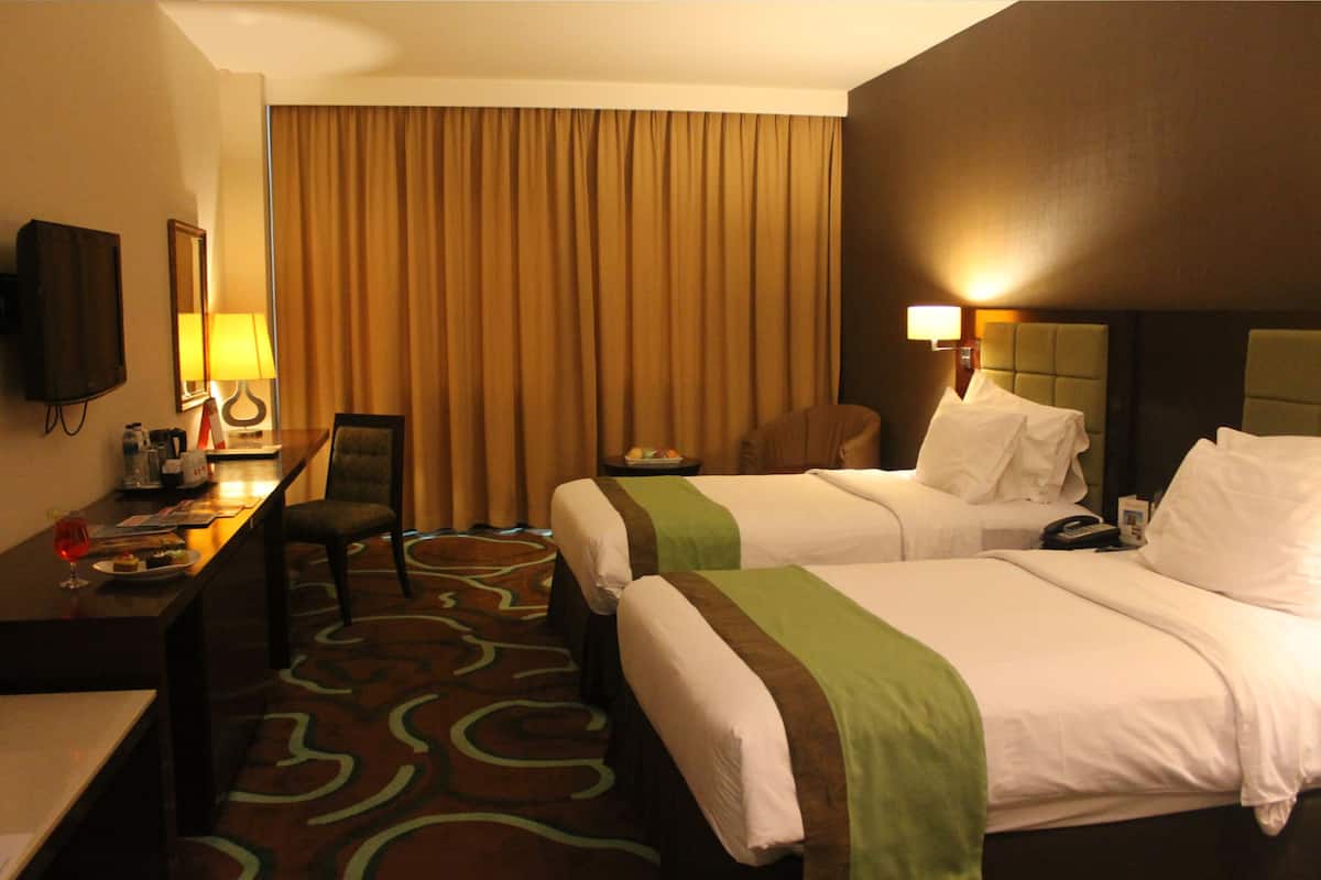 A hotel room with two beds, a desk with a chair, a television, and a large window with curtains.
