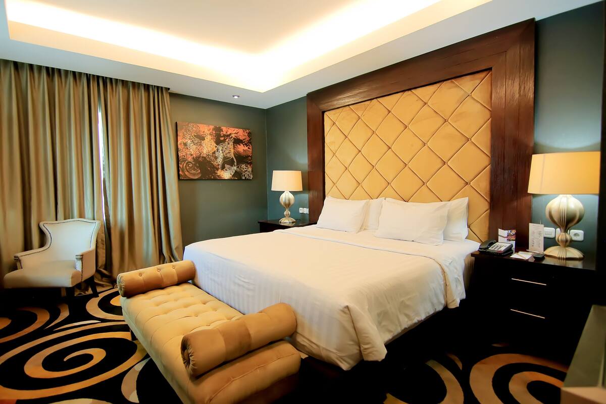 A hotel room with a large bed, a chair, a bench, a nightstand with a lamp, and a wall art piece.