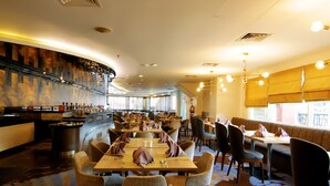 Un restaurante con varias mesas y sillas, una zona de bar y amplios ventanales con cortinas.