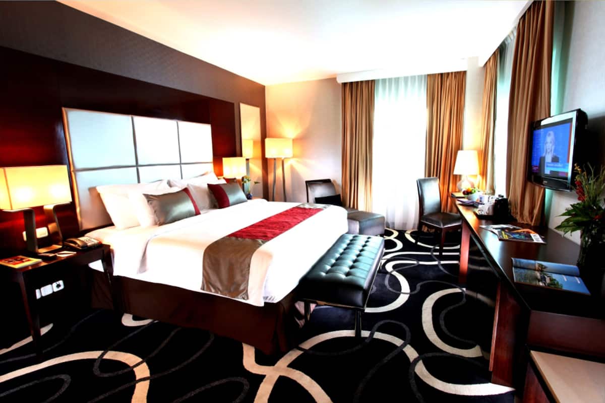 A hotel room with a large bed, bedside tables, a desk with a chair, a television, and a potted plant.