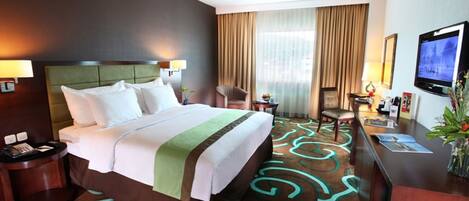 A hotel room with a large bed, a desk with a TV, a chair, and a window with curtains.