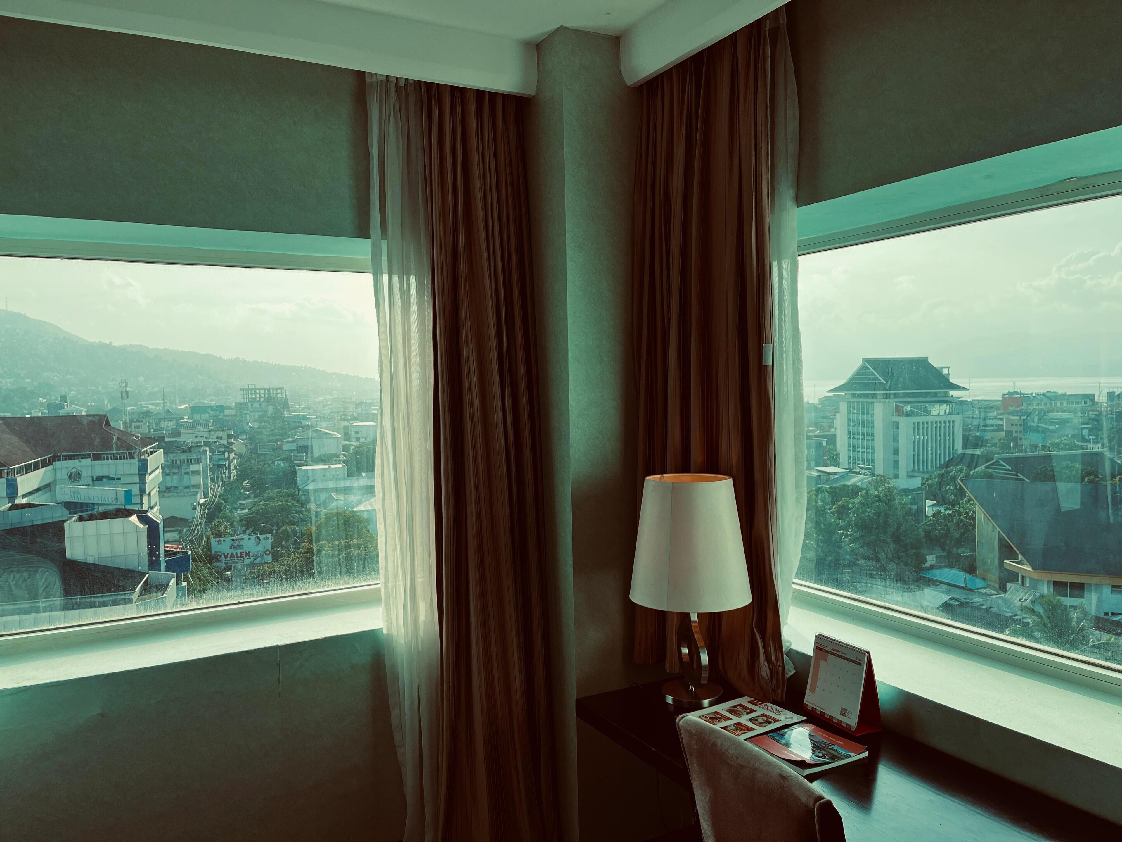 A hotel room with a large window, curtains, a desk with a lamp, and a calendar.