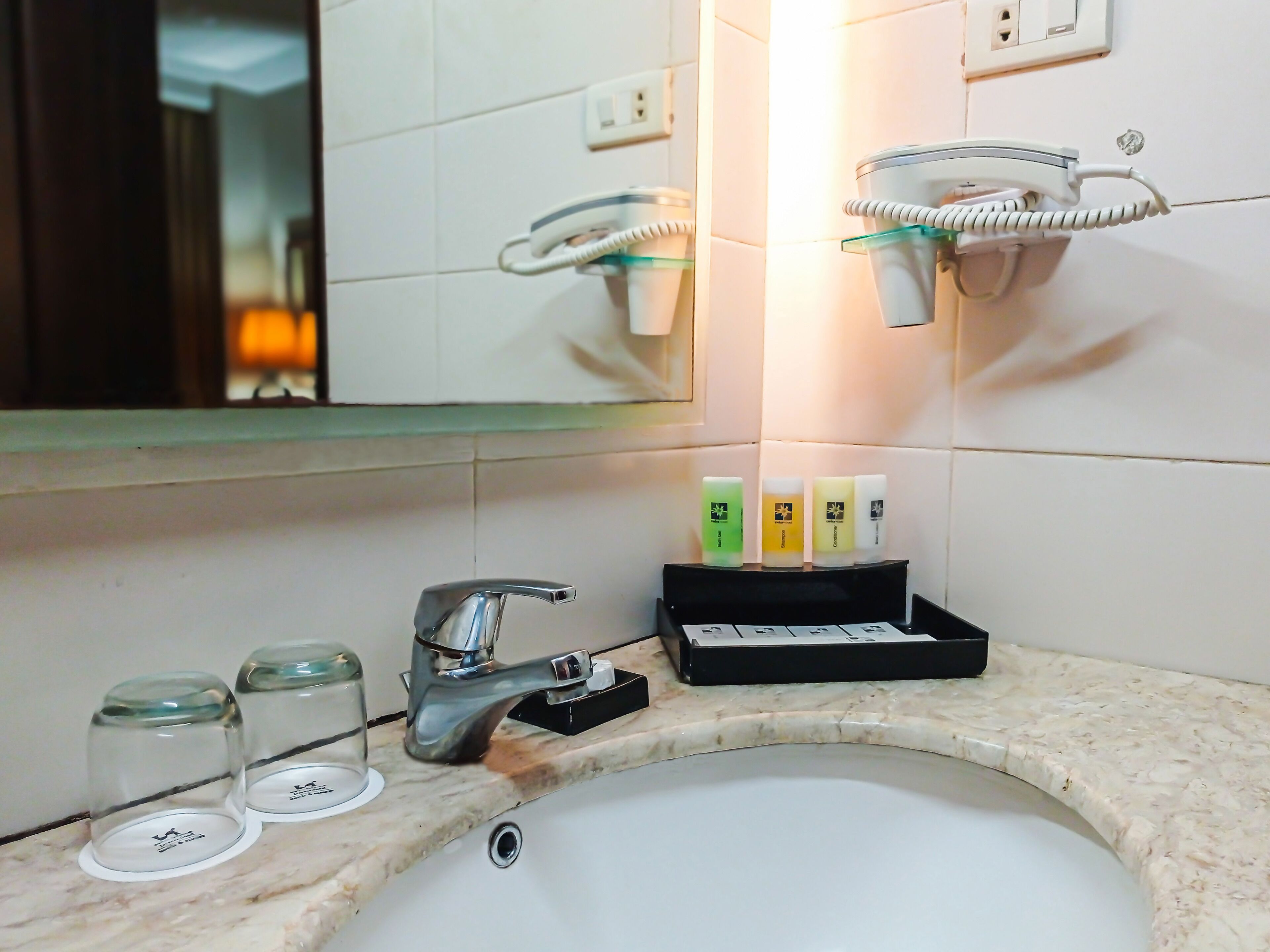 Une salle de bain avec un lavabo blanc, un miroir, un distributeur de savon en verre et un petit plateau noir contenant des articles de toilette.