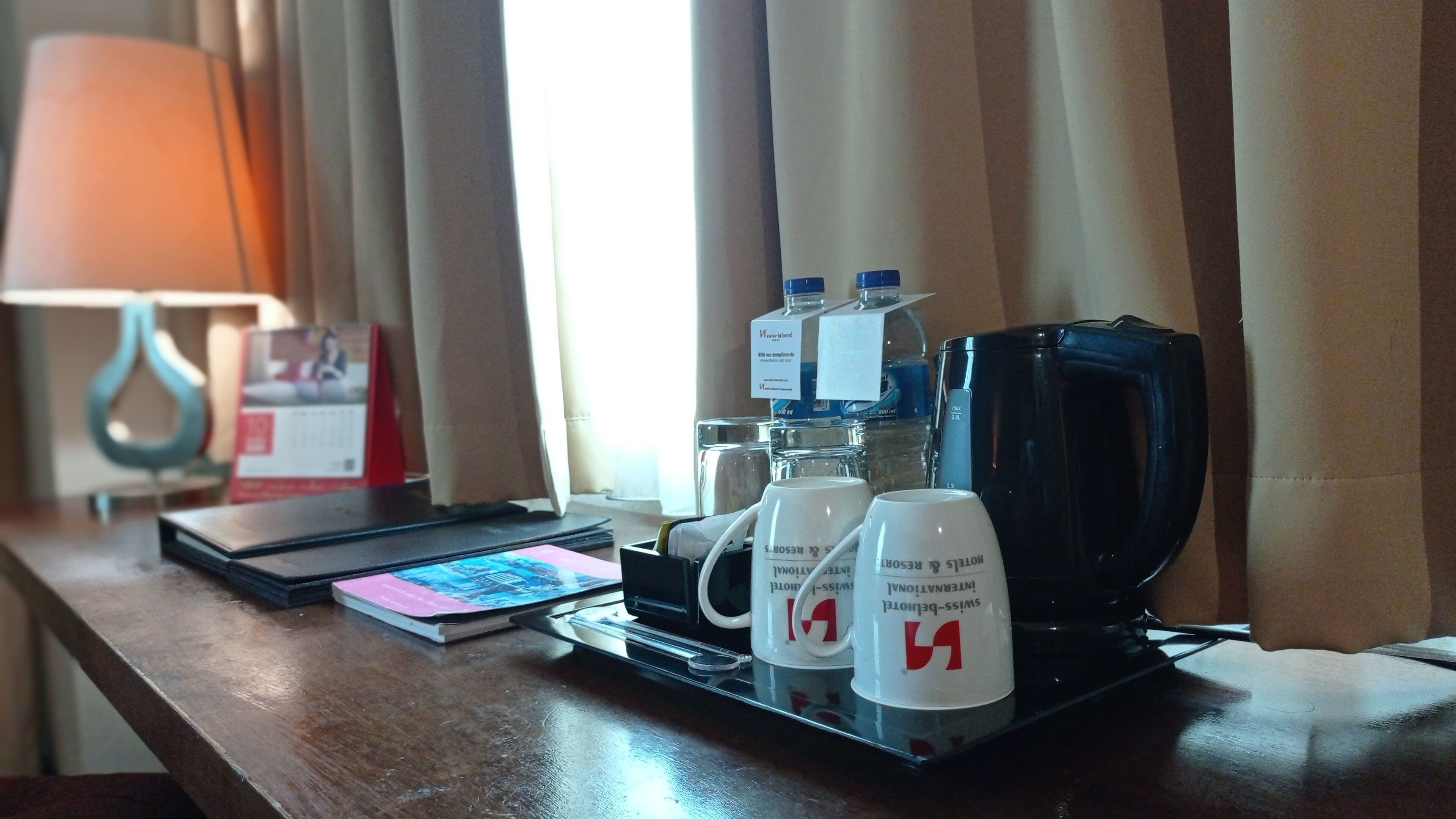 A hotel room with a coffee maker, two mugs, a notebook, and a lamp on a wooden table.