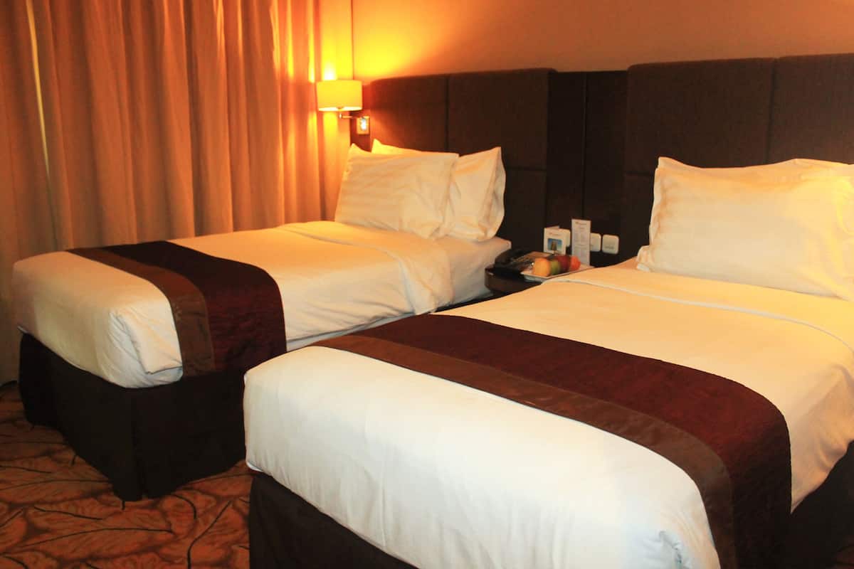 A hotel room with two beds, each with white linens and a brown accent. There is a nightstand with a lamp and a fruit bowl.
