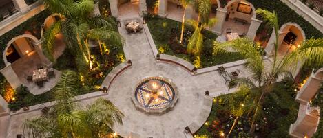 Un patio central con una fuente, rodeado de palmeras y zonas de estar, con senderos y arcos de piedra.