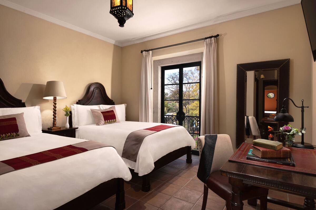 A hotel room with two beds, a desk, a chair, a window with curtains, a lamp, and a mirror.