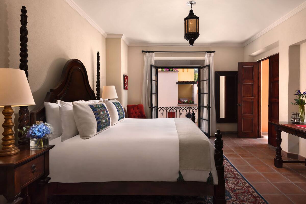 A neatly made bed with a white comforter and multiple pillows, a wooden nightstand with a lamp, and a view of a balcony through an open door.