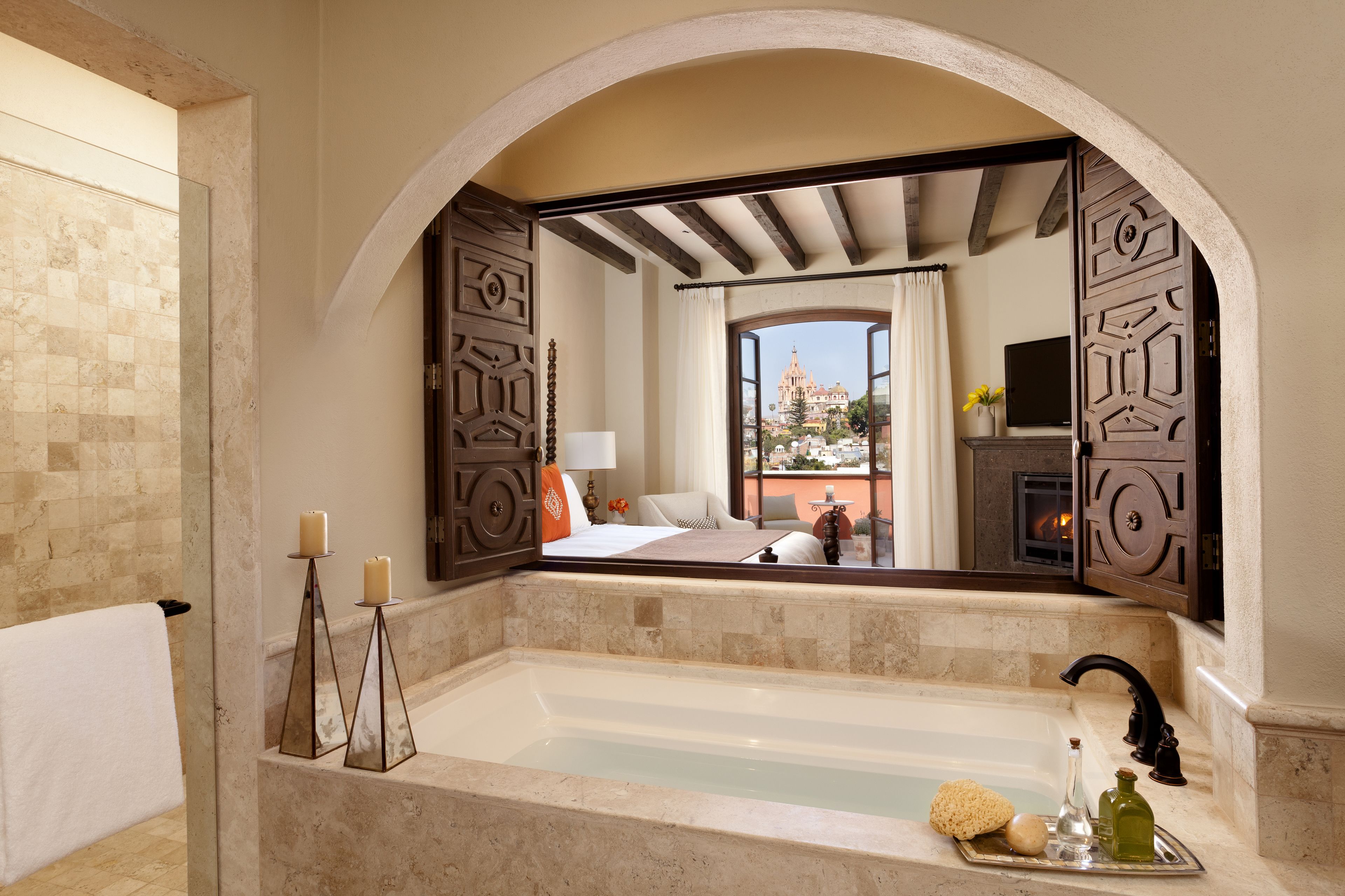 A bathroom with a large mirror, a bathtub, and a view of a bedroom with a bed and a TV.