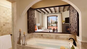 A bathroom with a large mirror, a bathtub, and a view of a bedroom with a bed and a TV.
