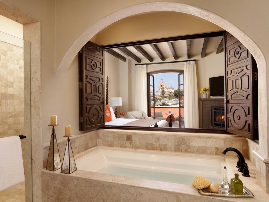 A bathroom with a large mirror, a bathtub, and a view of a bedroom with a bed and a TV.