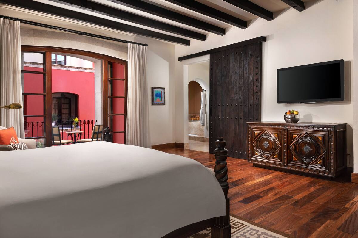 A hotel room with a large bed, a wooden dresser, a flat-screen TV, and a view of an outdoor area with a red wall and a balcony.