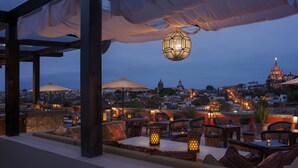 An outdoor seating area with tables and chairs, a hanging lantern, and a view of a cityscape with buildings and a church in the background. - Rosewood San Miguel De Allende (San Miguel de Allende)