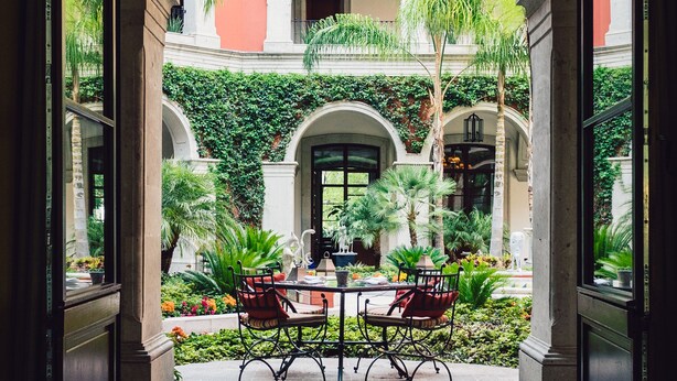 An open archway leading to a courtyard with a table and chairs, surrounded by greenery and a building with a red facade. - Rosewood San Miguel De Allende (San Miguel de Allende)