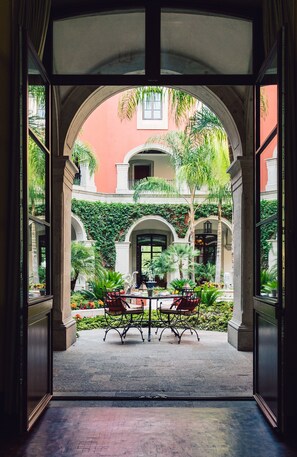 Un arco abierto que da a un patio con una mesa y sillas, rodeado de follaje y un edificio de fachada roja.