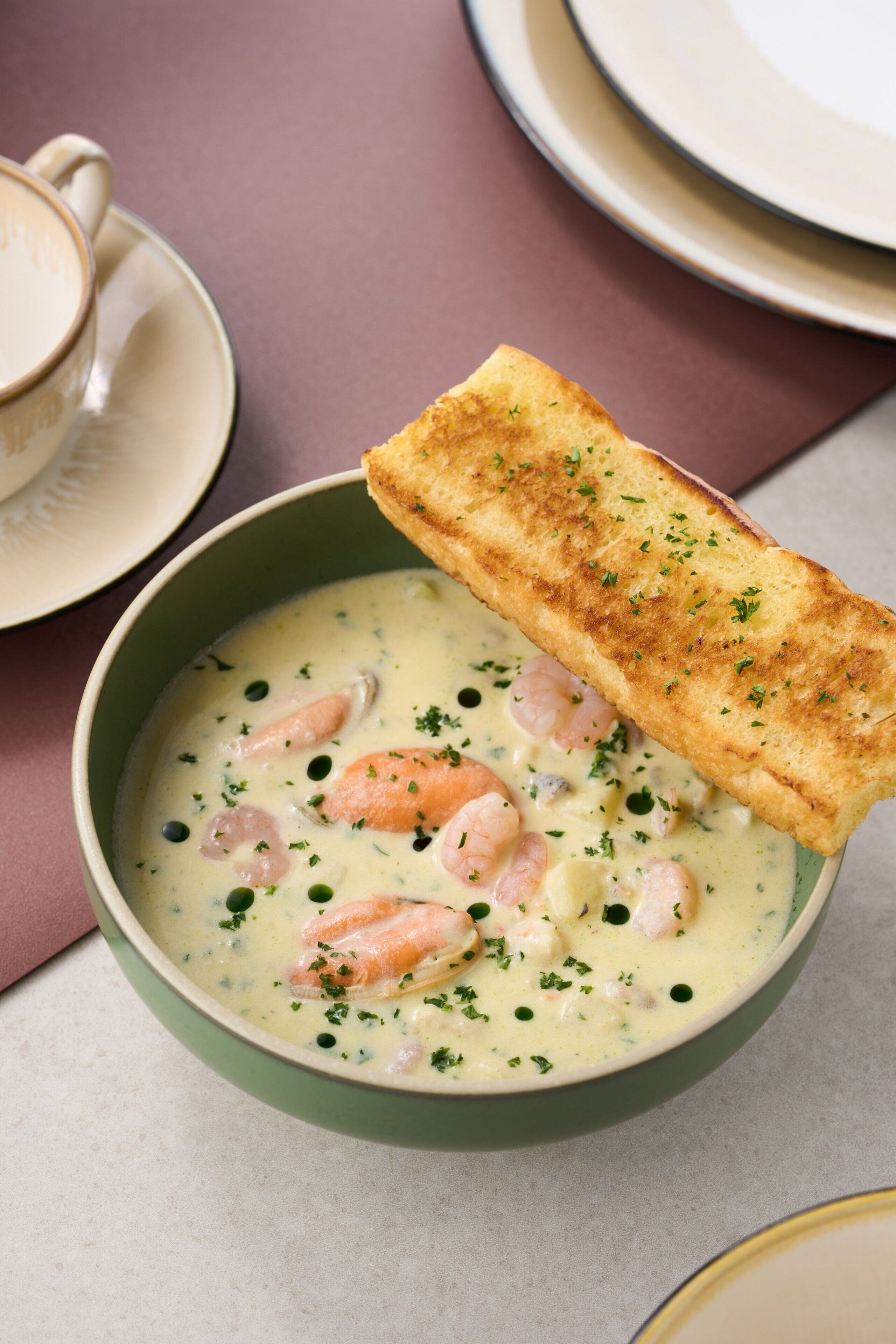 A bowl of creamy seafood soup garnished with herbs, accompanied by a toasted bread slice.