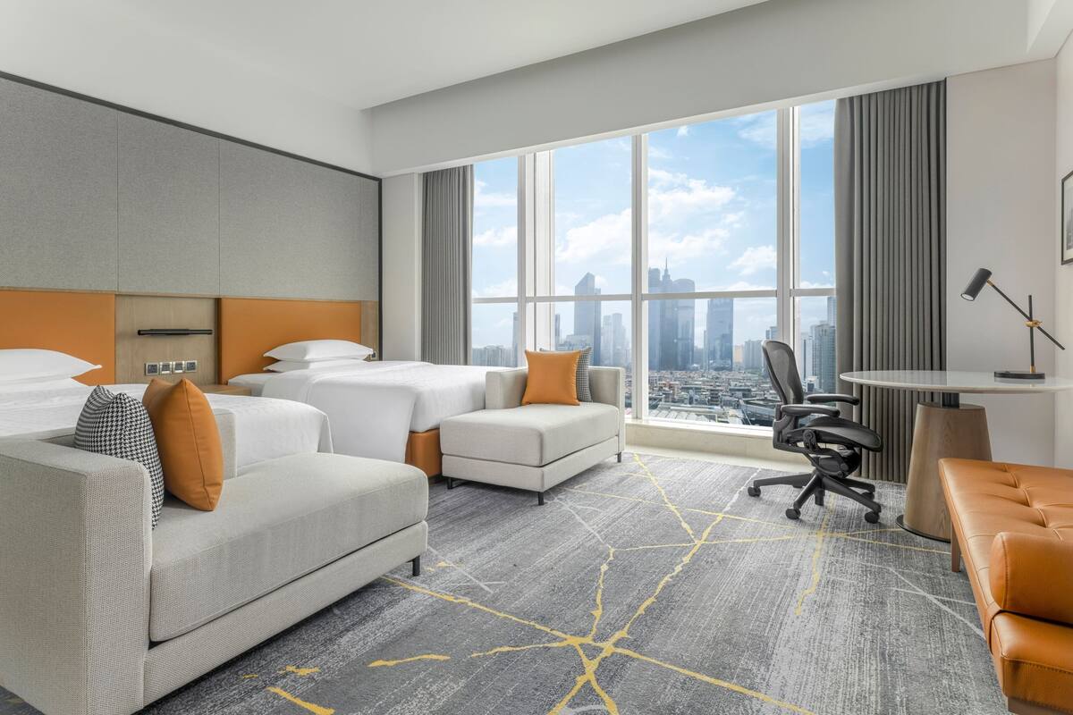 A hotel room with a large window, two beds, a sofa, a desk, and a chair.