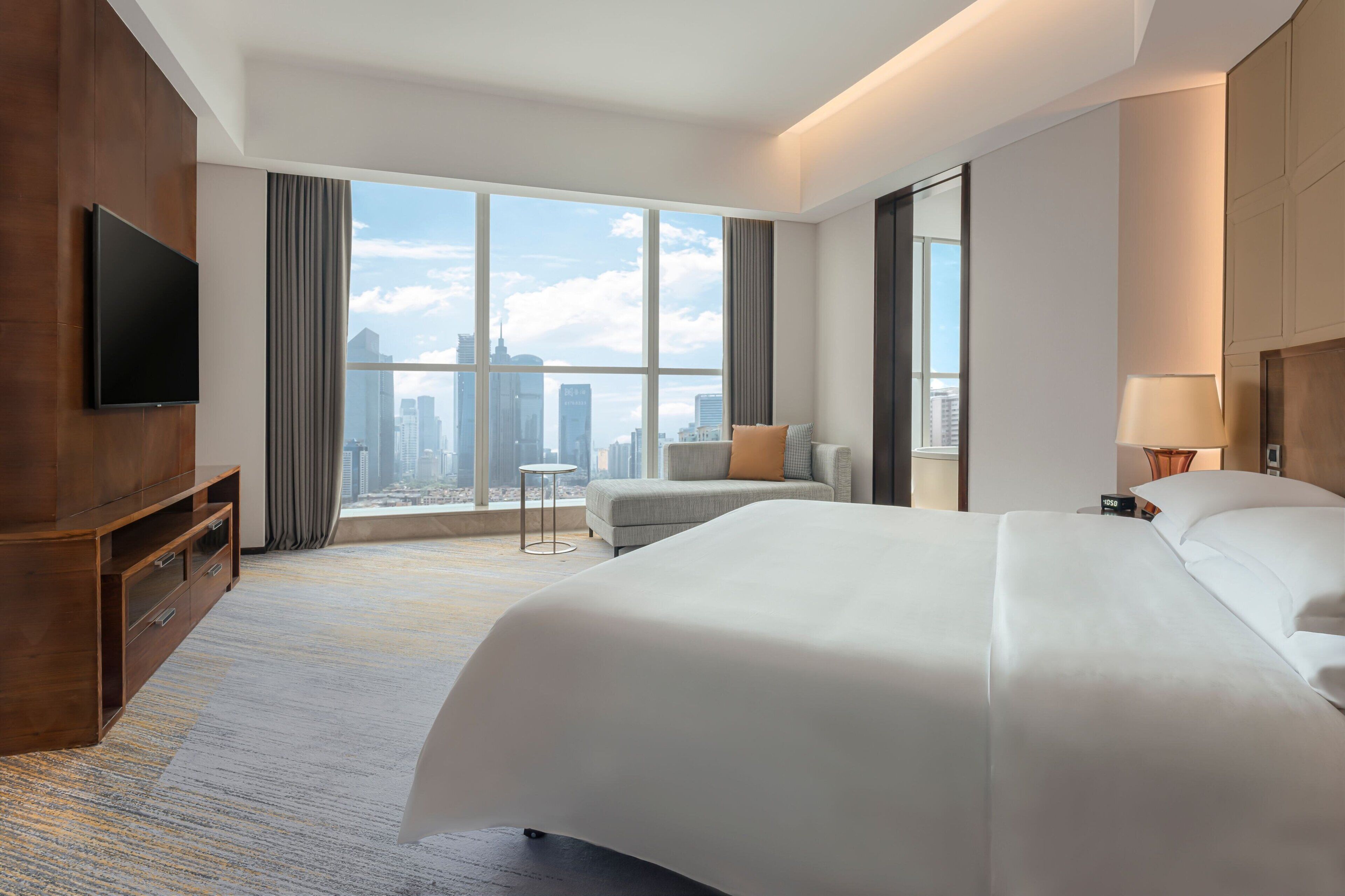 A hotel room with a large bed, a TV mounted on the wall, a city view through the window, and a small table by the window.