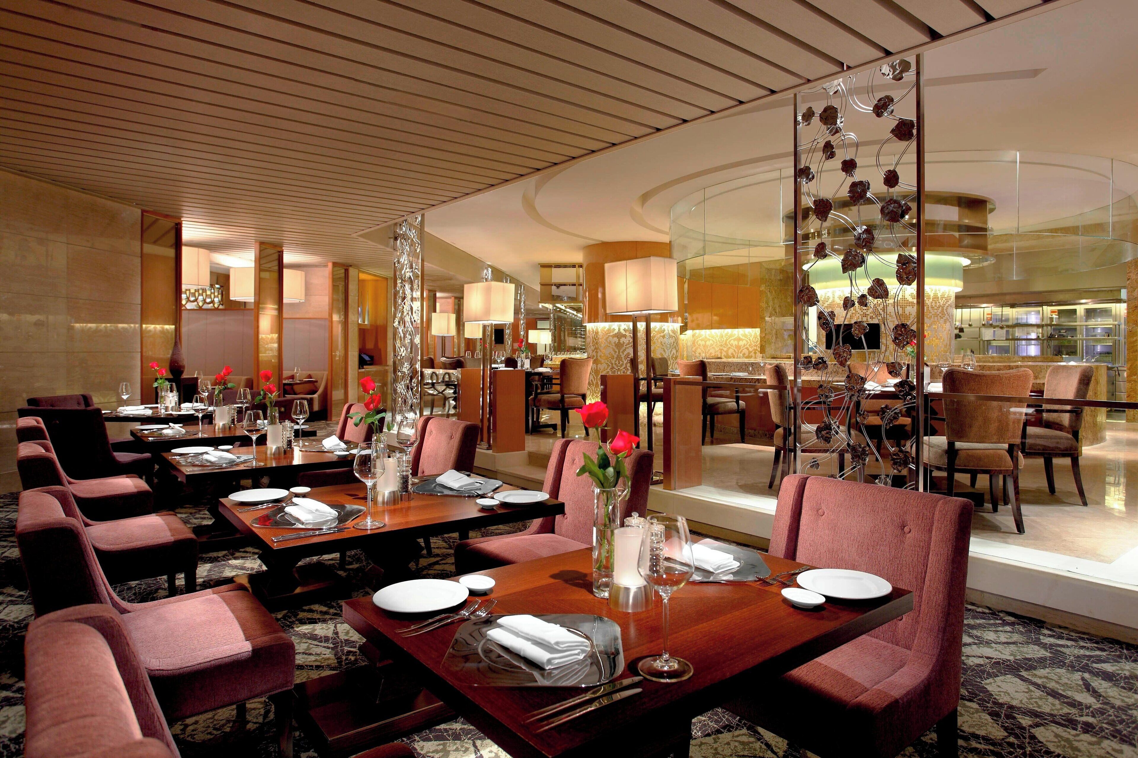 A hotel dining area with multiple tables set for dining, equipped with chairs, plates, cutlery, and a vase with flowers.