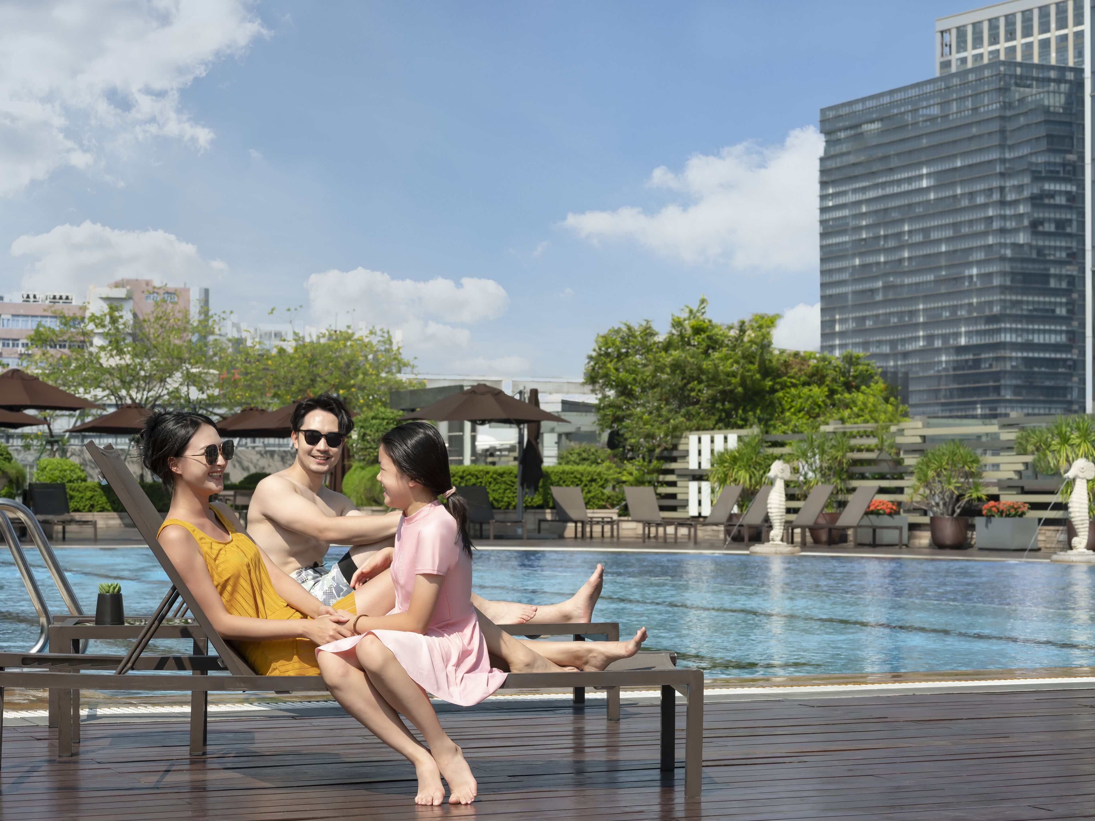 Zona junto a la piscina con sillones de descanso y vista a un edificio moderno al fondo.