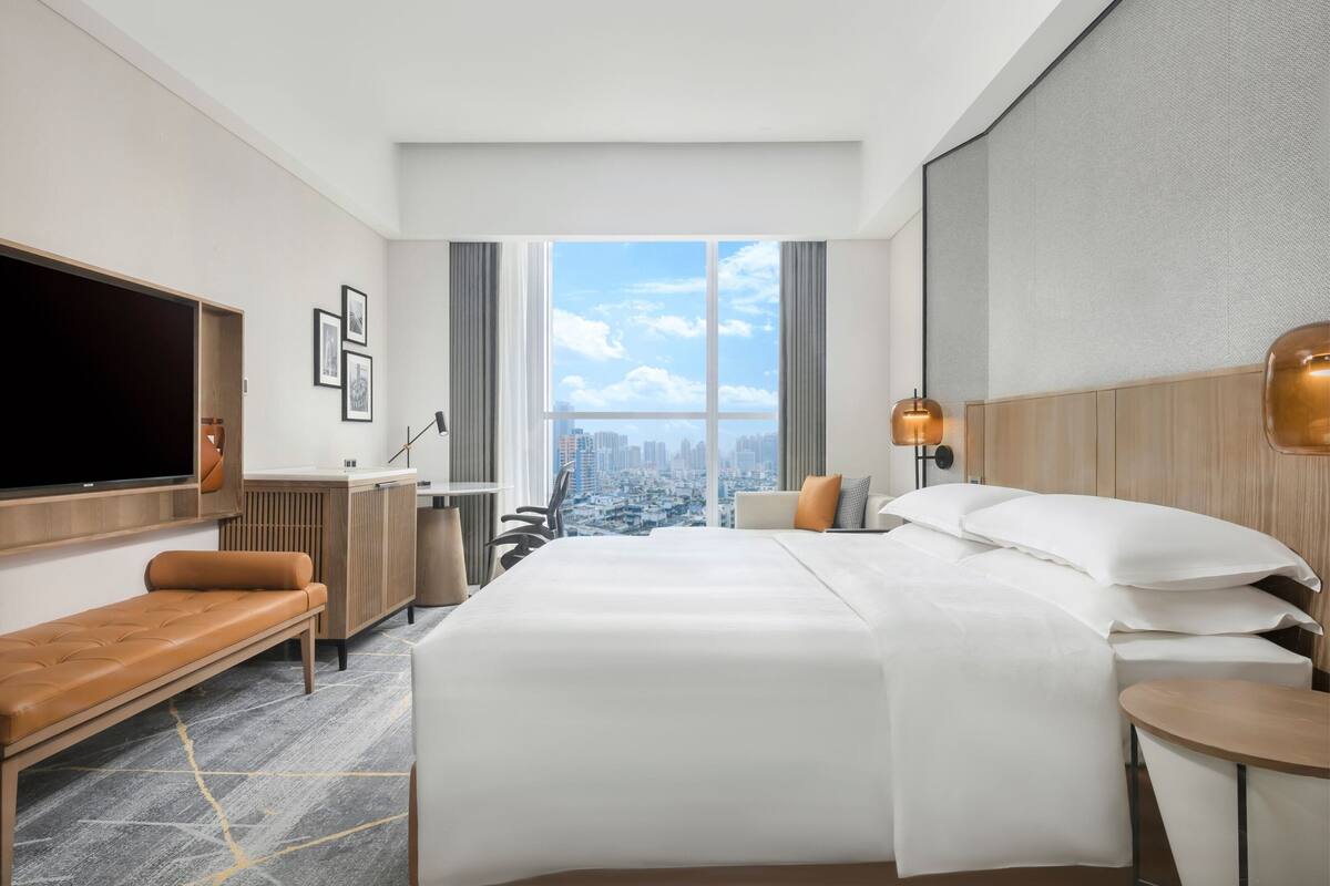 A modern hotel room with a large bed, a desk, a chair, a TV, and a city view.