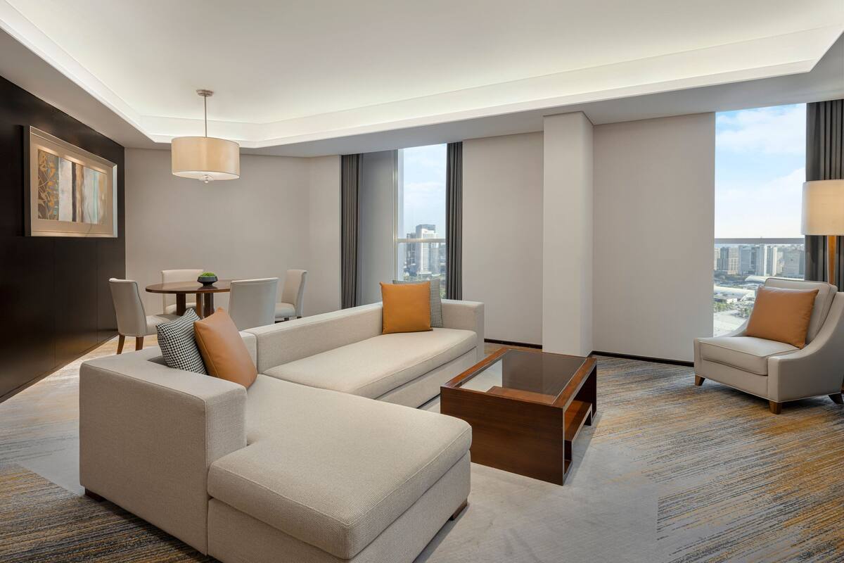 A modern hotel room with a sectional sofa, a small table, and a chair. There is a large window offering a city view.
