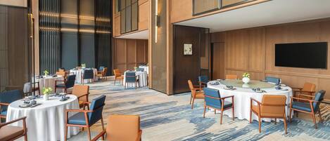 A modern hotel conference room with multiple round tables, chairs, and a large screen on the wall.
