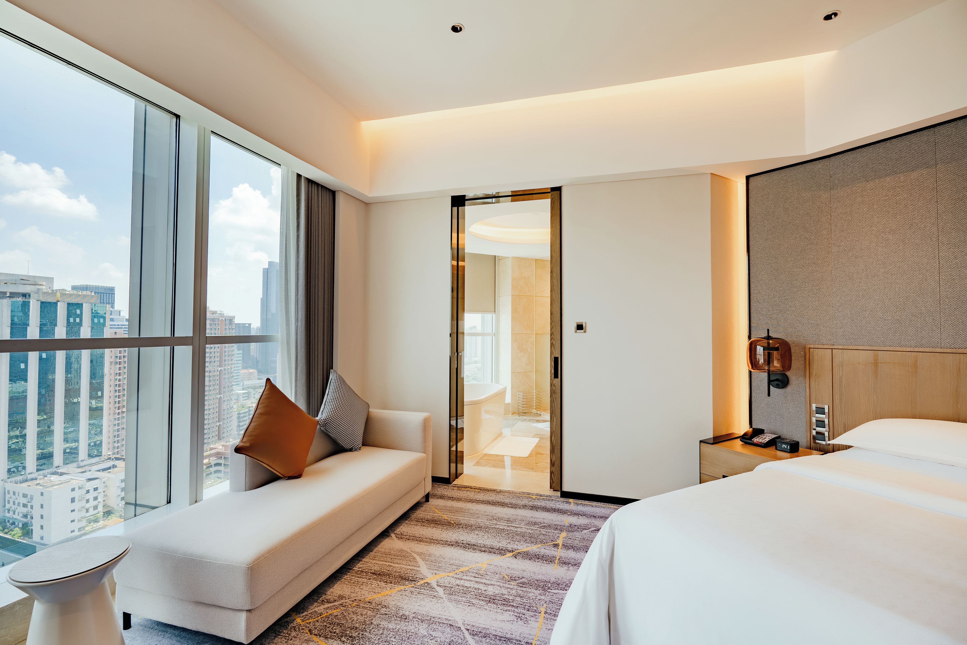 A modern hotel room with a large window, a sofa, a bed, a bedside table, and a bathroom visible through an open door.