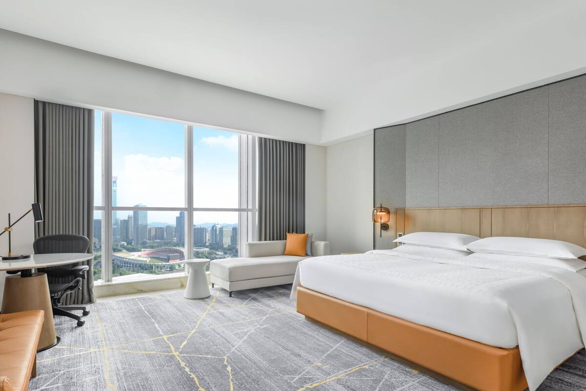 A modern hotel room with a large bed, a desk, a chair, and a sofa. There is a large window with a city view.