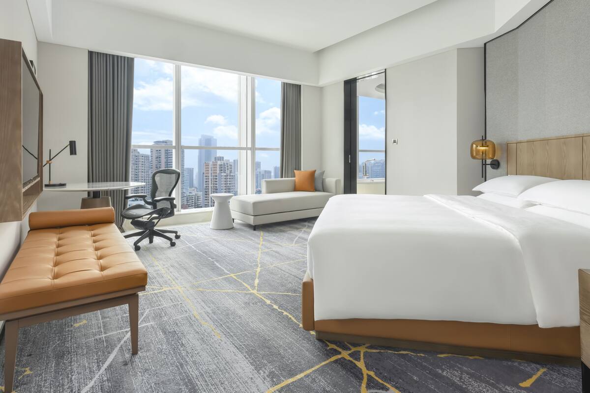 A modern hotel room with a large bed, a sofa, a desk, and a view of the cityscape through large windows.